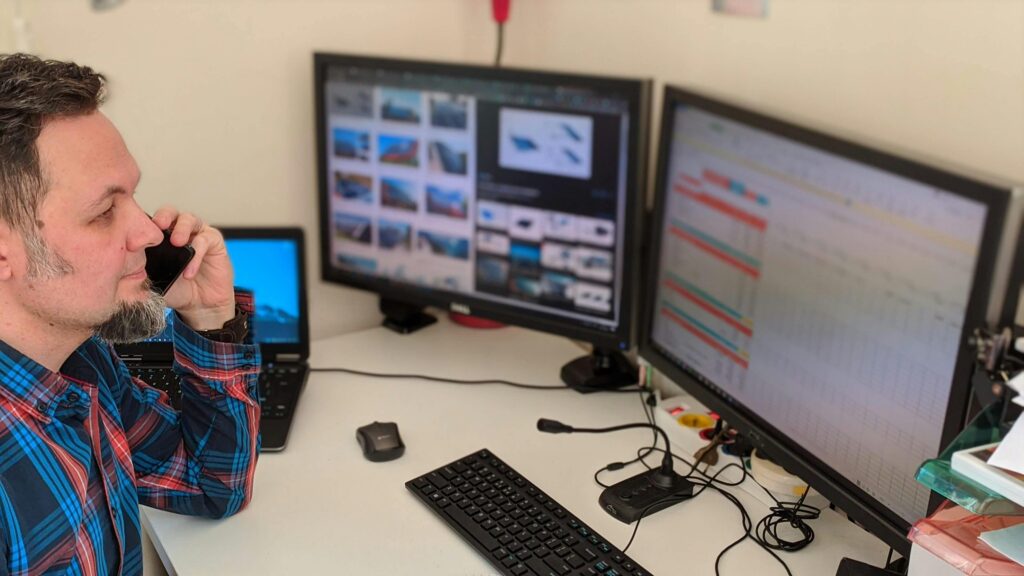 Mężczyzna w koszuli w kratę rozmawia przez telefon przy biurku z monitorami ze zdjęciami i arkuszem