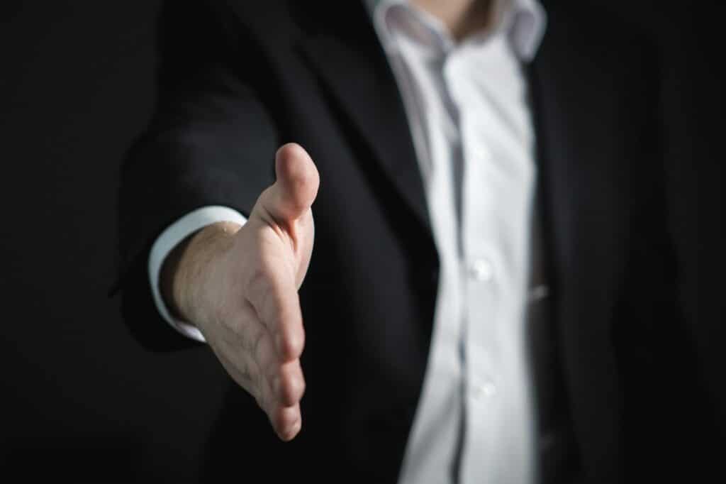 Mężczyzna w ciemnym garniturze i białej koszuli wyciąga dłoń na powitanie