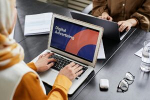 Kobieta w chuście pisze na laptopie ze slajdem Digital Advertising, przy stole siedzi koleżanka