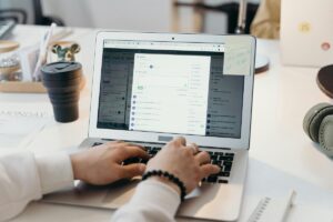 Dłonie piszące na laptopie z tablicą zadań, a na biurku kubek kawy, słuchawki i notes