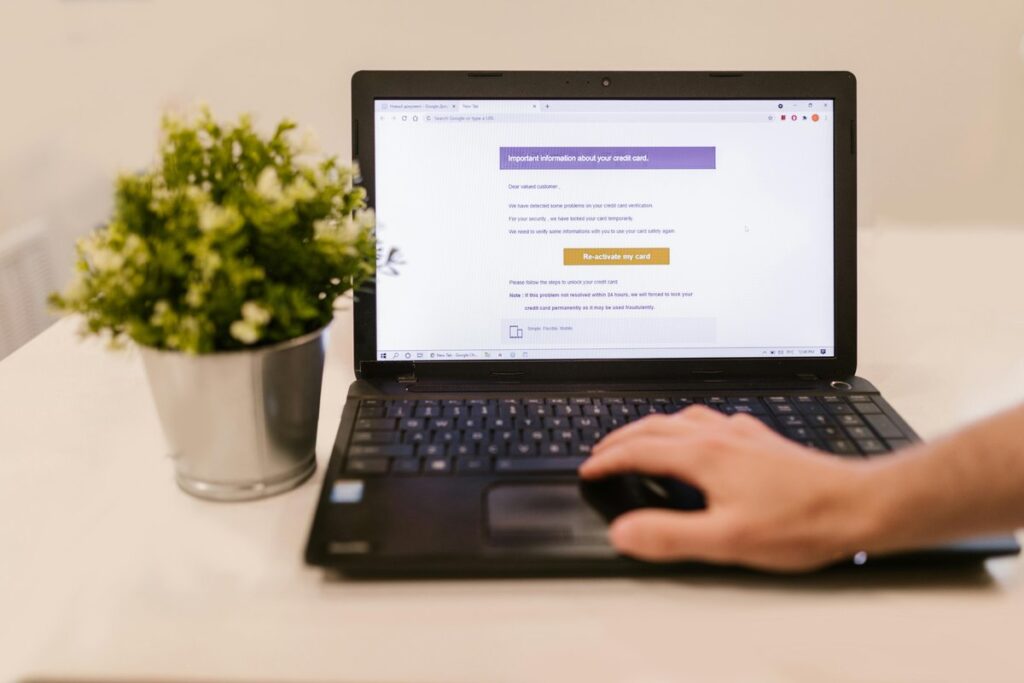 Dłoń na myszce przy laptopie z podejrzanym e-mailem o karcie kredytowej, obok roślina w doniczce