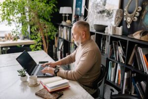 Gray-bearded man in a beige sweater working on a laptop at a marble desk beside bookshelves